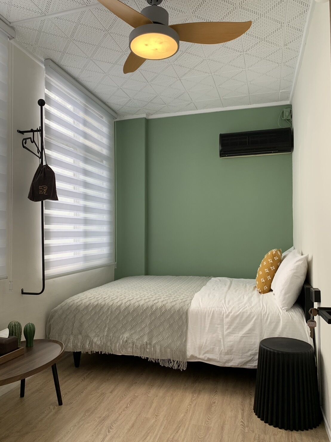 Un dormitorio con cama, ventilador de techo, ventana con persianas, una mesita y un calentador fijado a la pared.