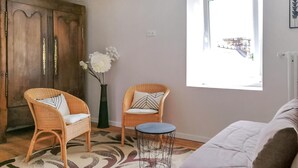 Una habitación con dos sillones de mimbre, una mesita redonda pequeña, un sofá blanco, un tapete estampado, un armario de madera y una ventana con vistas.