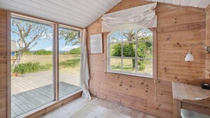 Una cabaña de madera con un amplio ventanal corredera que da a un espacio exterior, una ventana pequeña con vistas a la vegetación y un sencillo escritorio de madera con una lámpara.