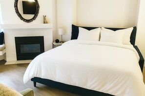 A neatly arranged bedroom with a large bed, a fireplace, a small table with a lamp, and a wall-mounted TV.