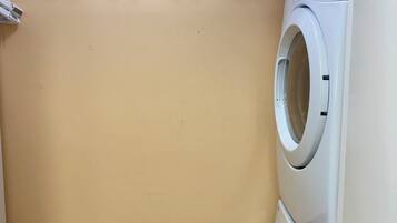 Une buanderie équipée d’un lave-linge et d’un sèche-linge superposés, d’une étagère avec des bouteilles et d’un panier contenant de la lessive.