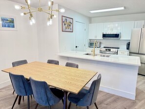 A modern kitchen with white cabinets, a stainless steel refrigerator, a microwave, and a stove. There is a wooden dining table with blue chairs.