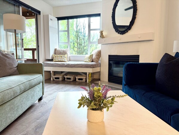 A living room with a sofa, a blue couch, a coffee table with a plant, a fireplace, and a window with a view of trees.