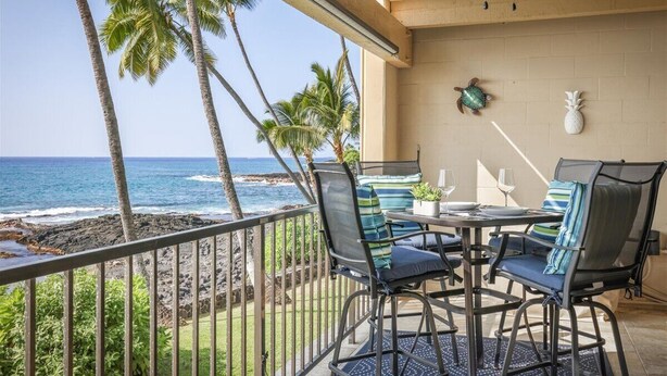 A balcony with a view of the ocean, featuring a table and chairs, a ceiling fan, and decorative wall art. - Direct Ocean Front Luxury Condo (Kailua-Kona)
