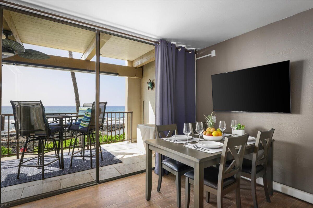 A dining area with a table set for four, a flat-screen TV mounted on the wall, and a view of the ocean through a sliding glass door.