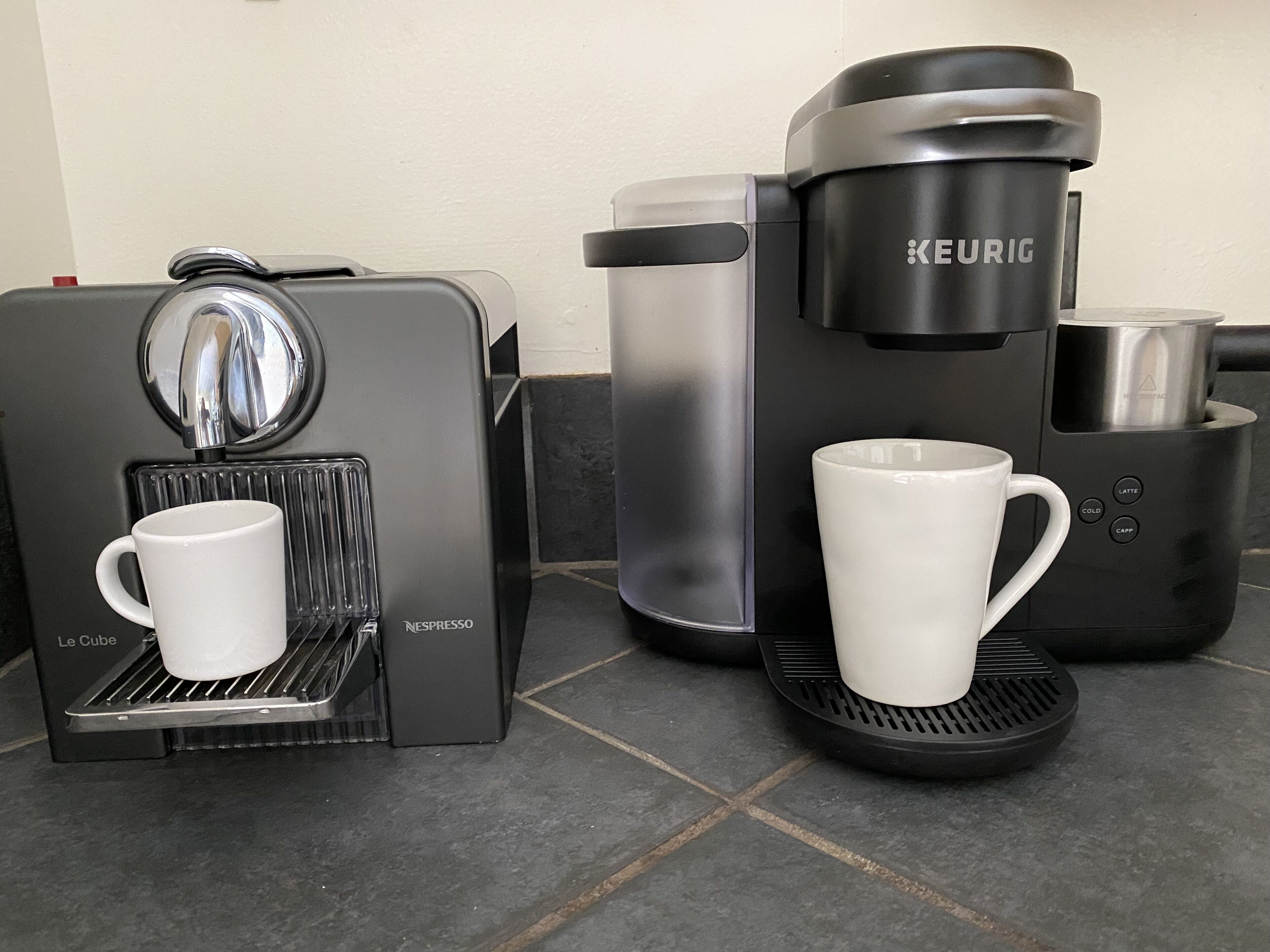 A Nespresso coffee machine with a white mug on the drip tray and a Keurig coffee maker with a white mug on the drip tray.
