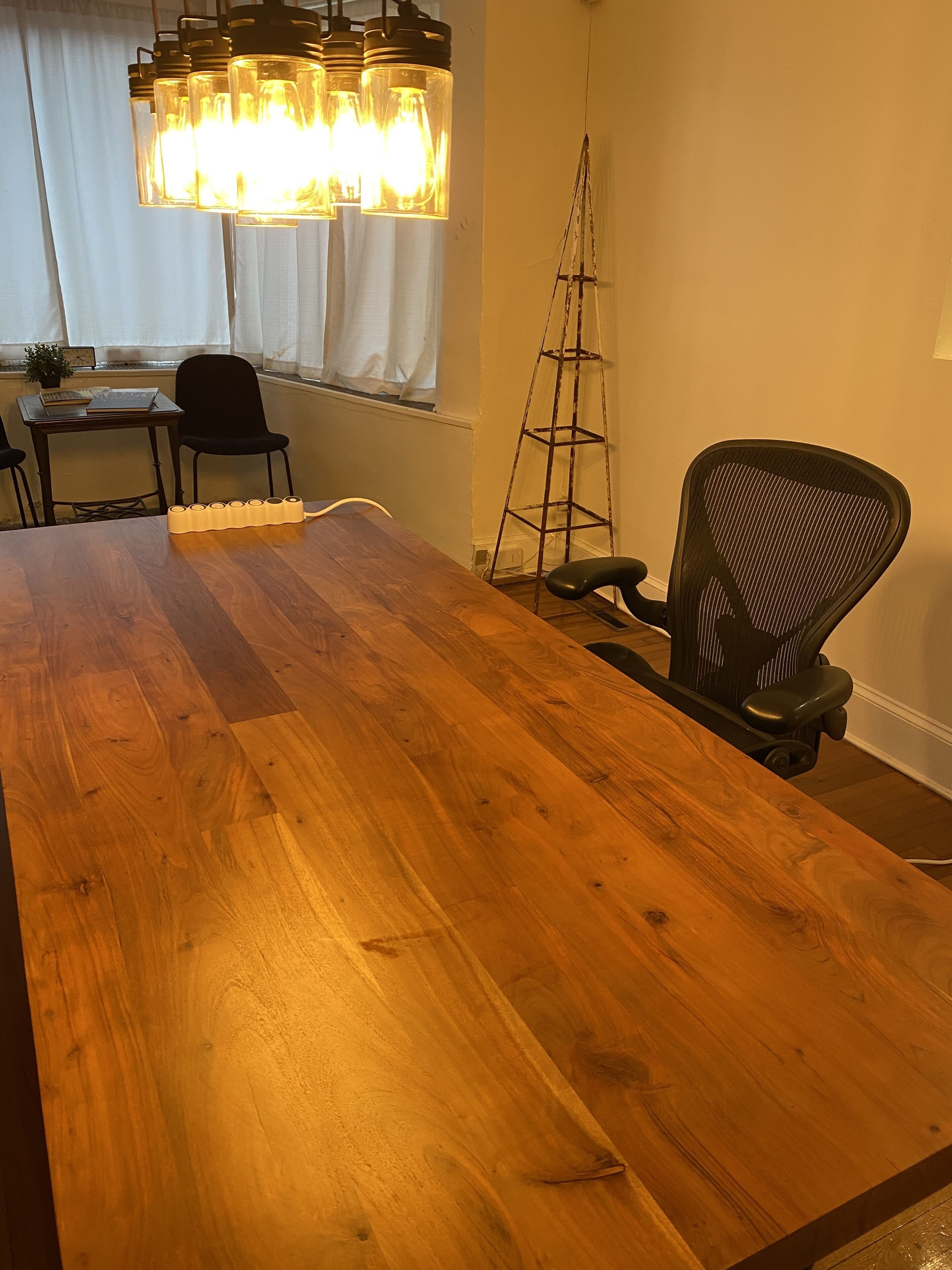 A wooden dining table with a black office chair, a lamp with exposed bulbs, and a ladder against the wall.