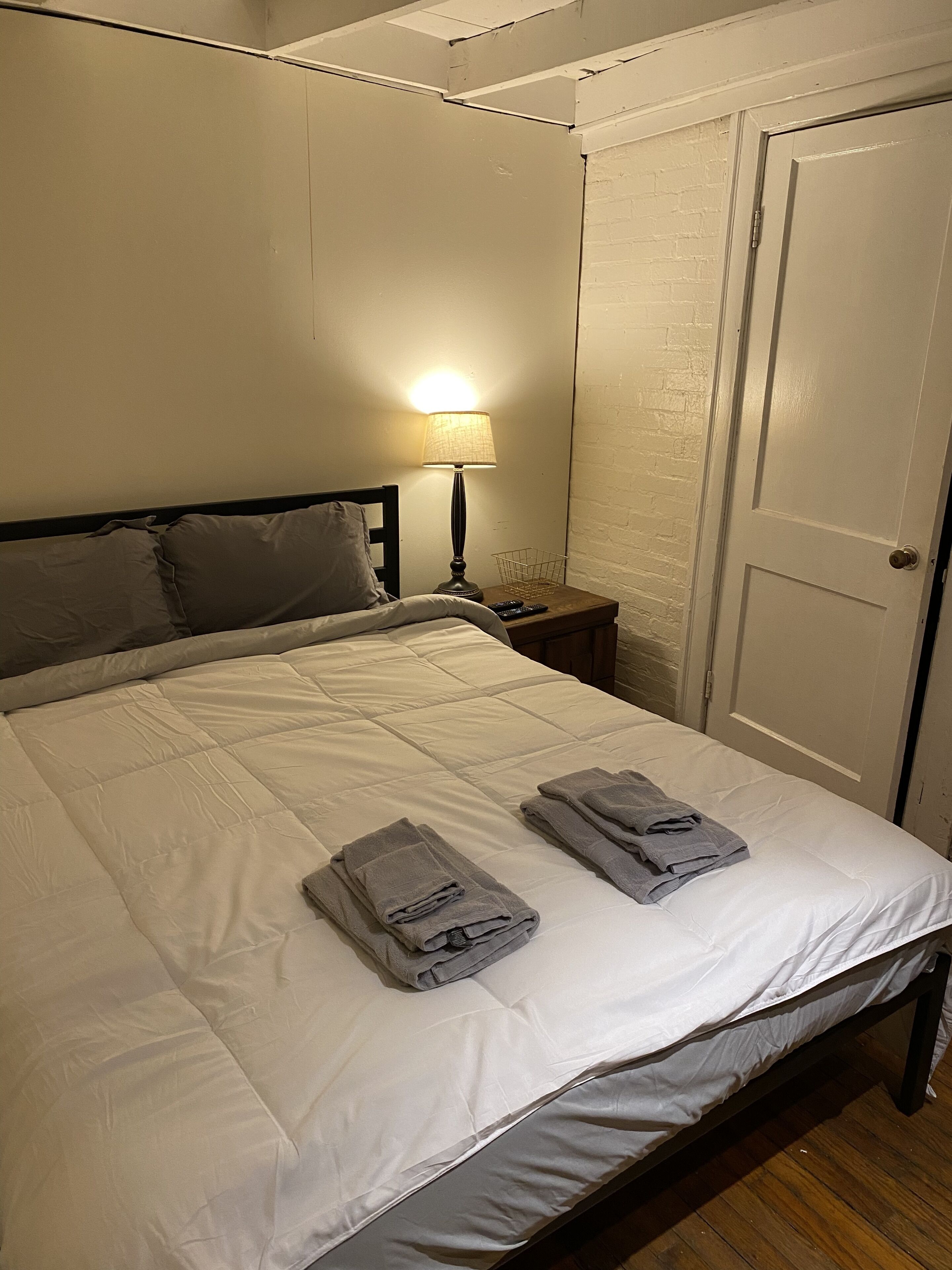 Ein ordentlich bezogenes Bett mit gefalteten Handtüchern, einer Nachttischlampe und einer geschlossenen Tür in einem Raum mit weißen Wänden und Holzböden.