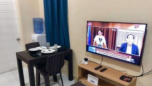 Ein Zimmer mit Fernseher, der eine Nachrichtensendung zeigt, einem Esstisch mit Geschirr und Besteck, einem Wasserspender und einem kleinen Holzregal mit elektronischen Geräten.