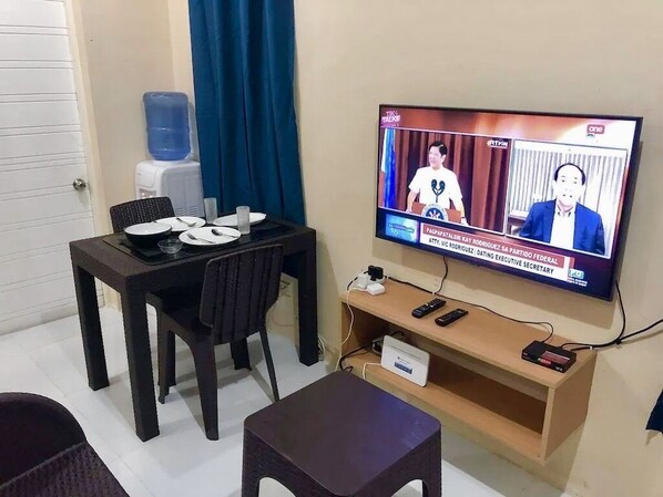Una habitación con una televisión mostrando un noticiero, una mesa de comedor con platos y cubiertos, un dispensador de agua y un pequeño estante de madera con dispositivos electrónicos.