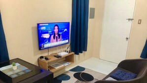 Ein Wohnzimmer mit Flachbildfernseher an der Wand, einer Couch, einem kleinen Tisch mit Pflanze und einem gemusterten Teppich.