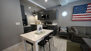 A modern kitchen with dark cabinets, a white countertop, and a dining area with a table and chairs. An American flag is displayed on the wall.