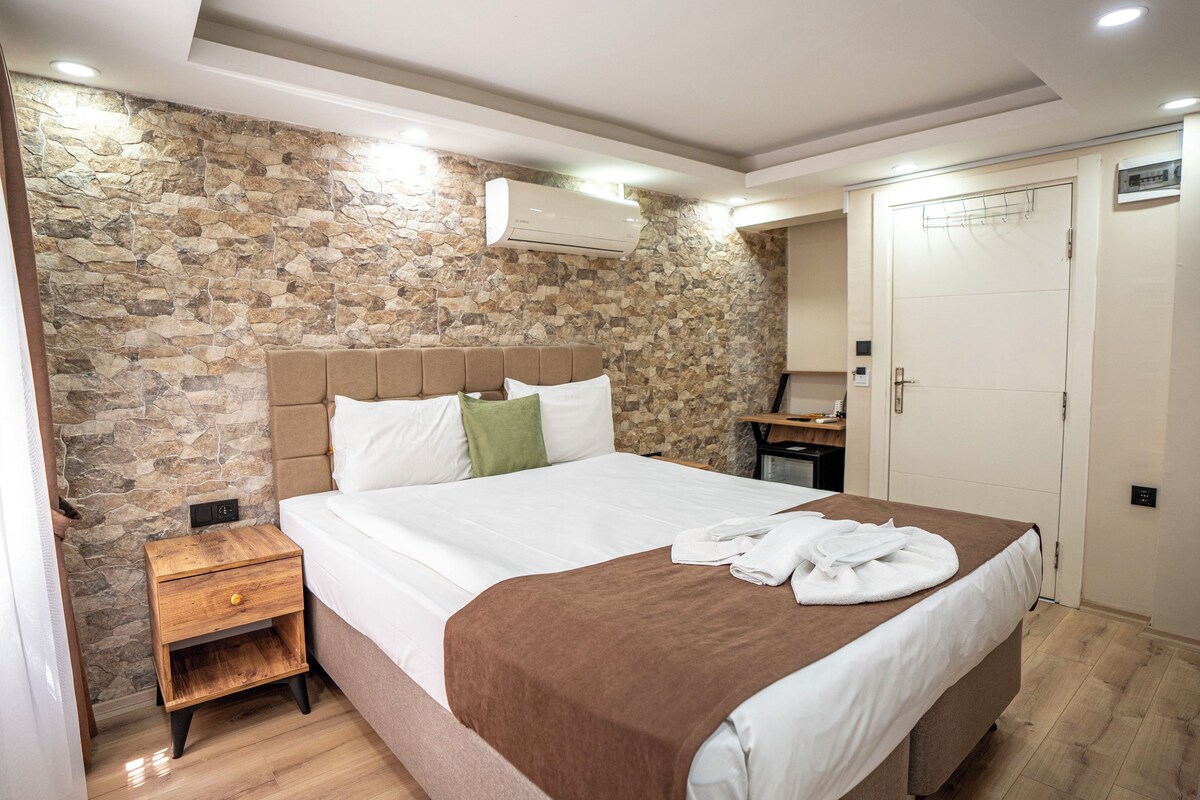 A bedroom with a stone wall, a bed with a brown headboard, a bedside table, a desk, and a wardrobe.
