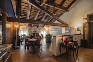A spacious dining area with wooden flooring, a high ceiling with exposed beams, and a view of the adjacent kitchen.