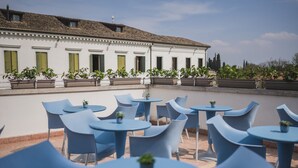 Zona salotto all'aperto con sedie blu e tavoli rotondi, circondata da piante in vaso e un edificio bianco con persiane verdi.