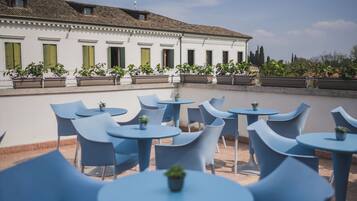 An outdoor seating area with blue chairs and tables, potted plants, and a building with green shutters in the background.