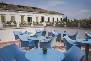 Een terras met blauwe stoelen en ronde tafels, omringd door potplanten en een wit gebouw met groene luiken.