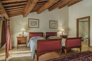 Een slaapkamer met een houten plafond, twee bedden met rode hoofdborden, bedlampen, een houten nachtkastje, een rood-wit gestreept gordijn en een kamerkleed met patroon.