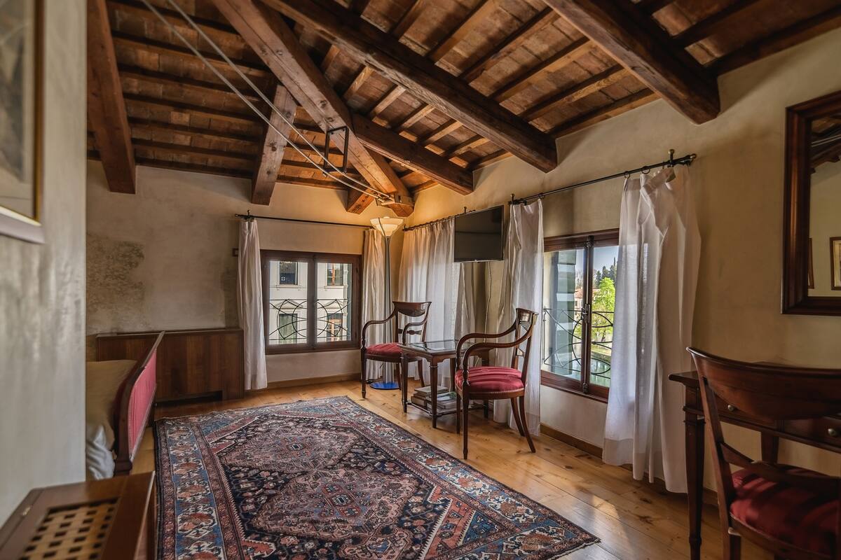 Ein Zimmer mit Holzbalken, einem großen Teppich, zwei Stühlen, einem Tisch und Blick durch ein Fenster.
