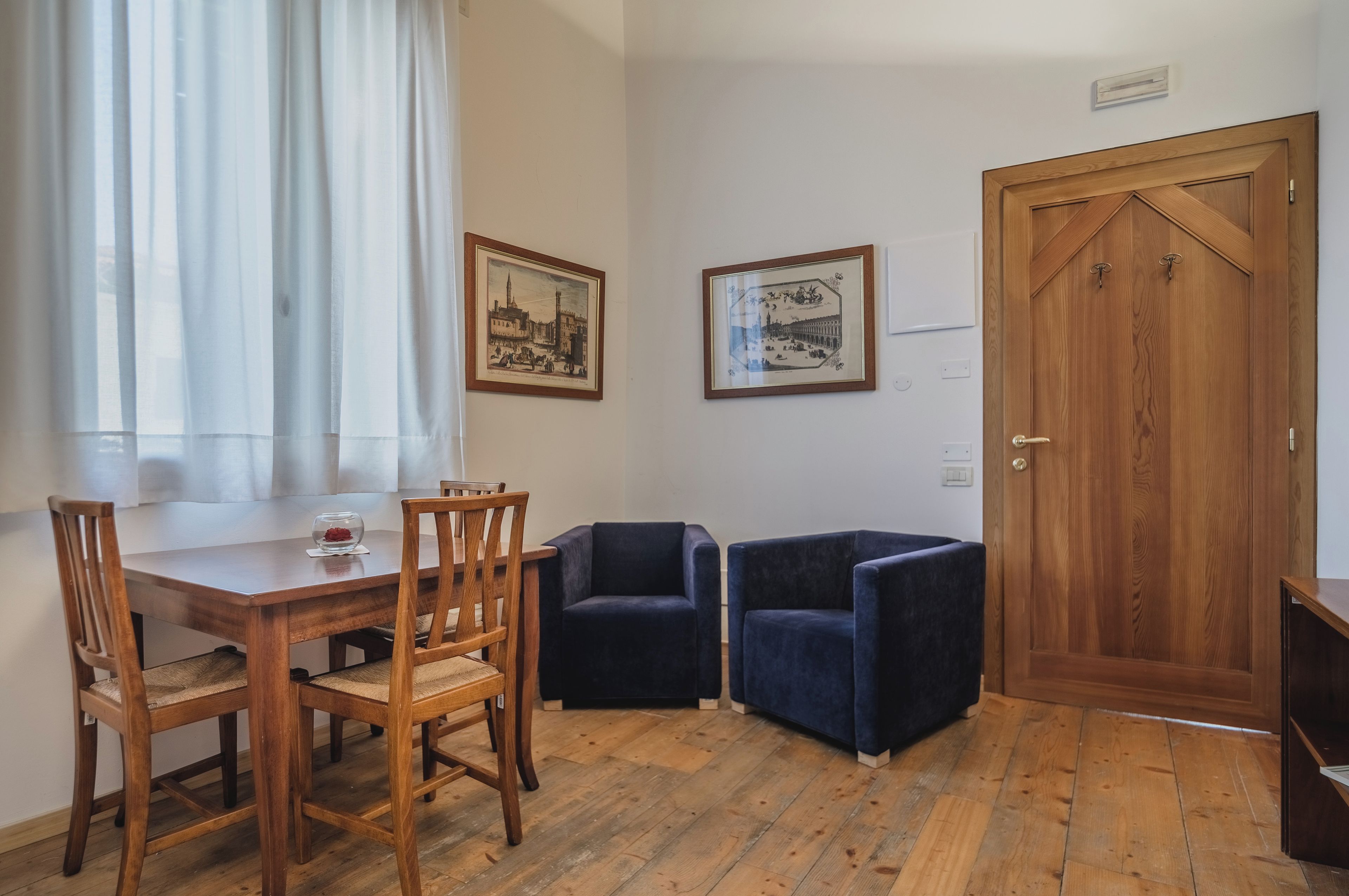 A room with a wooden dining table, two chairs, a wooden door, and two framed pictures on the wall.