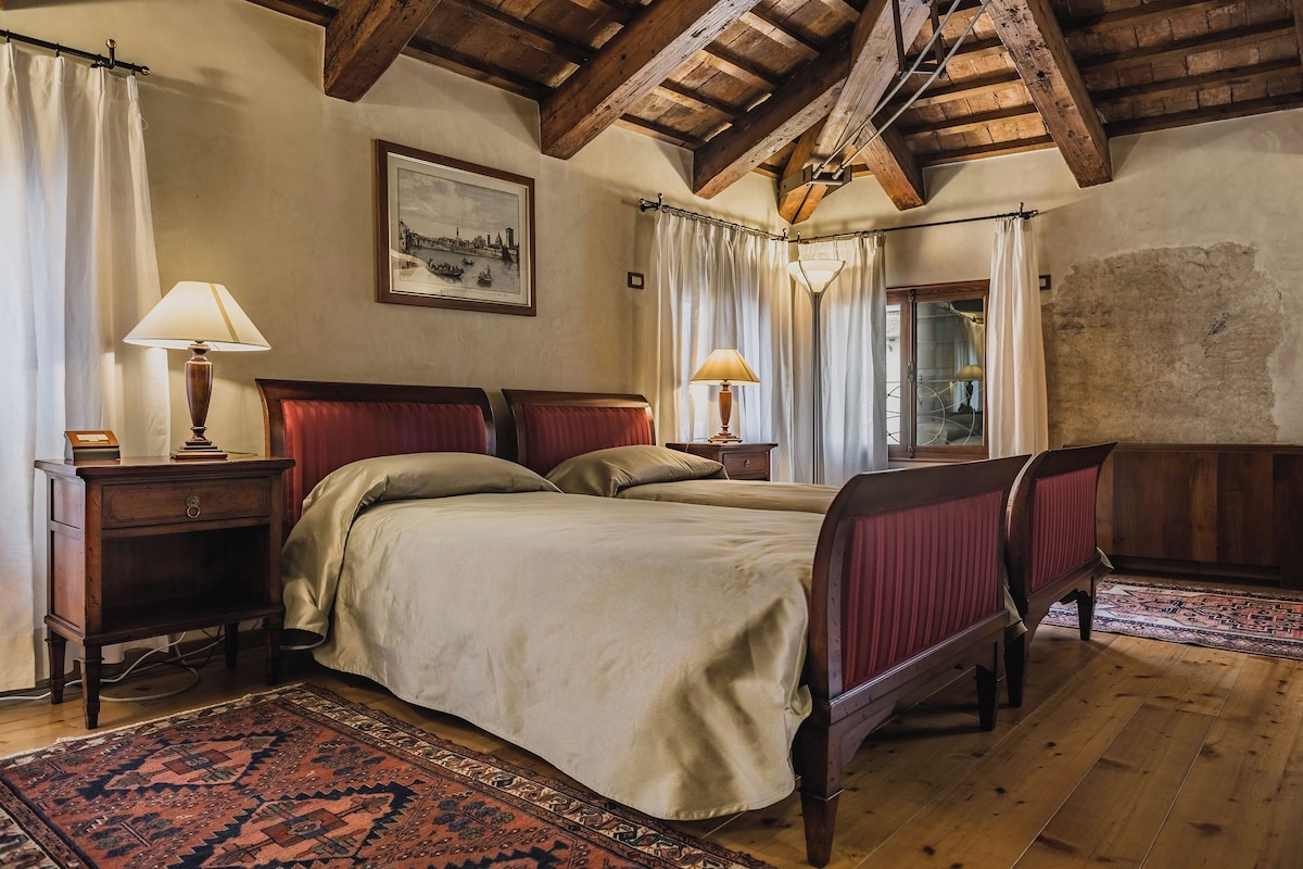 Une chambre à coucher avec deux lits, des têtes de lit en bois, une table de chevet, une lampe, un tableau et une grande fenêtre avec des rideaux.