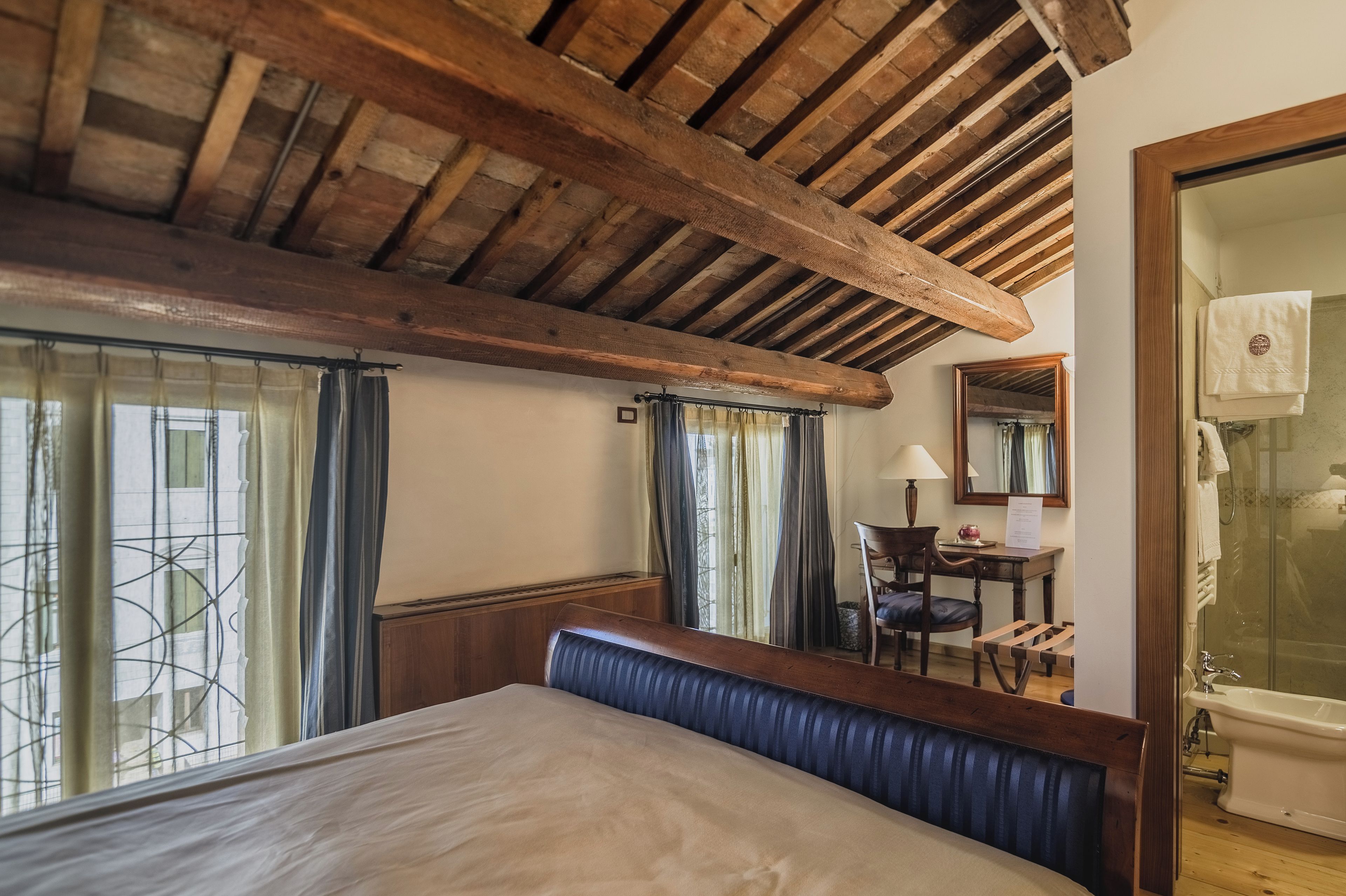 A bedroom with a wooden ceiling, a bed with a blue headboard, a desk with a chair, a mirror, and a bathroom with a bathtub.