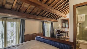 A bedroom with a wooden ceiling, a bed with a blue headboard, a desk with a chair, a mirror, and a bathroom with a bathtub.