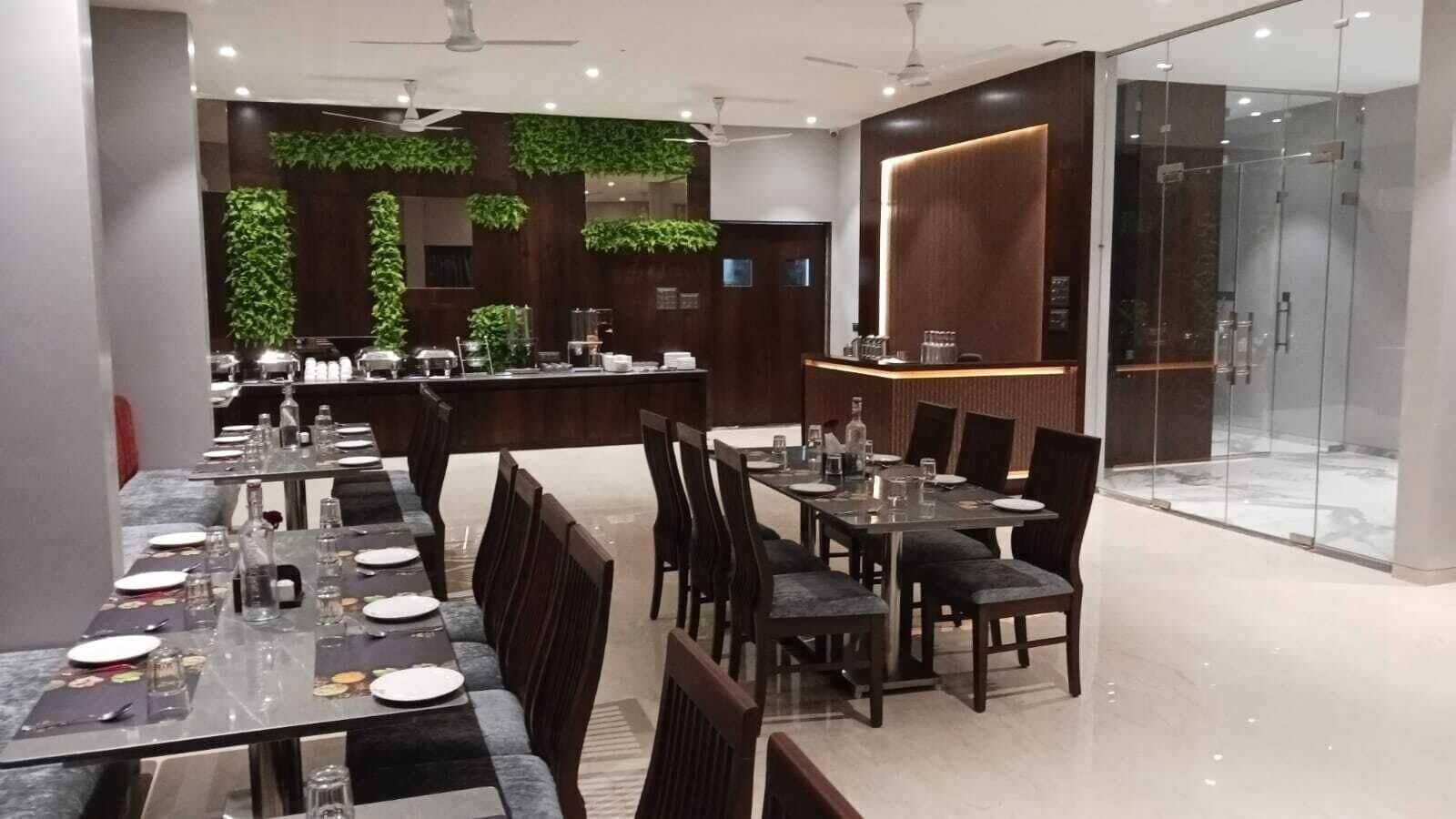 A modern restaurant with long tables, dark wooden chairs, and a bar area with a glass partition.