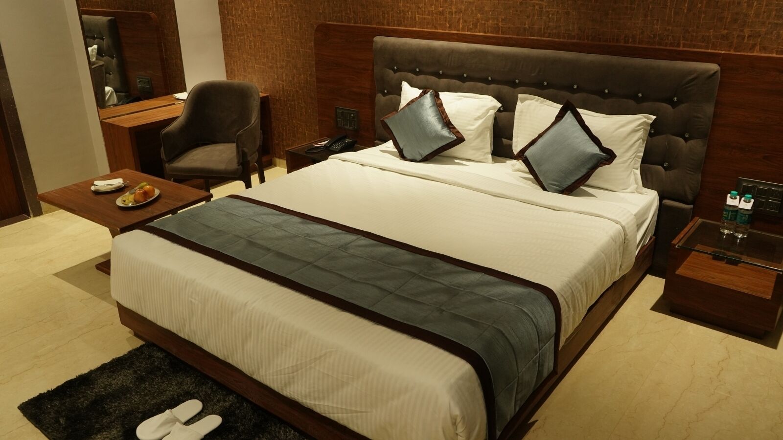A hotel room with a large bed, a wooden nightstand, a chair, and a small table with a plate of fruit.