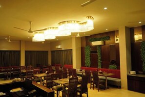 A hotel dining area with multiple tables and chairs, a ceiling fan, and decorative lighting fixtures. - Alaukik Hotel (Rahata)