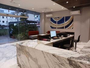 A modern reception desk with a marble counter, a laptop, and a computer monitor. There is a glass wall with a view of buildings outside.