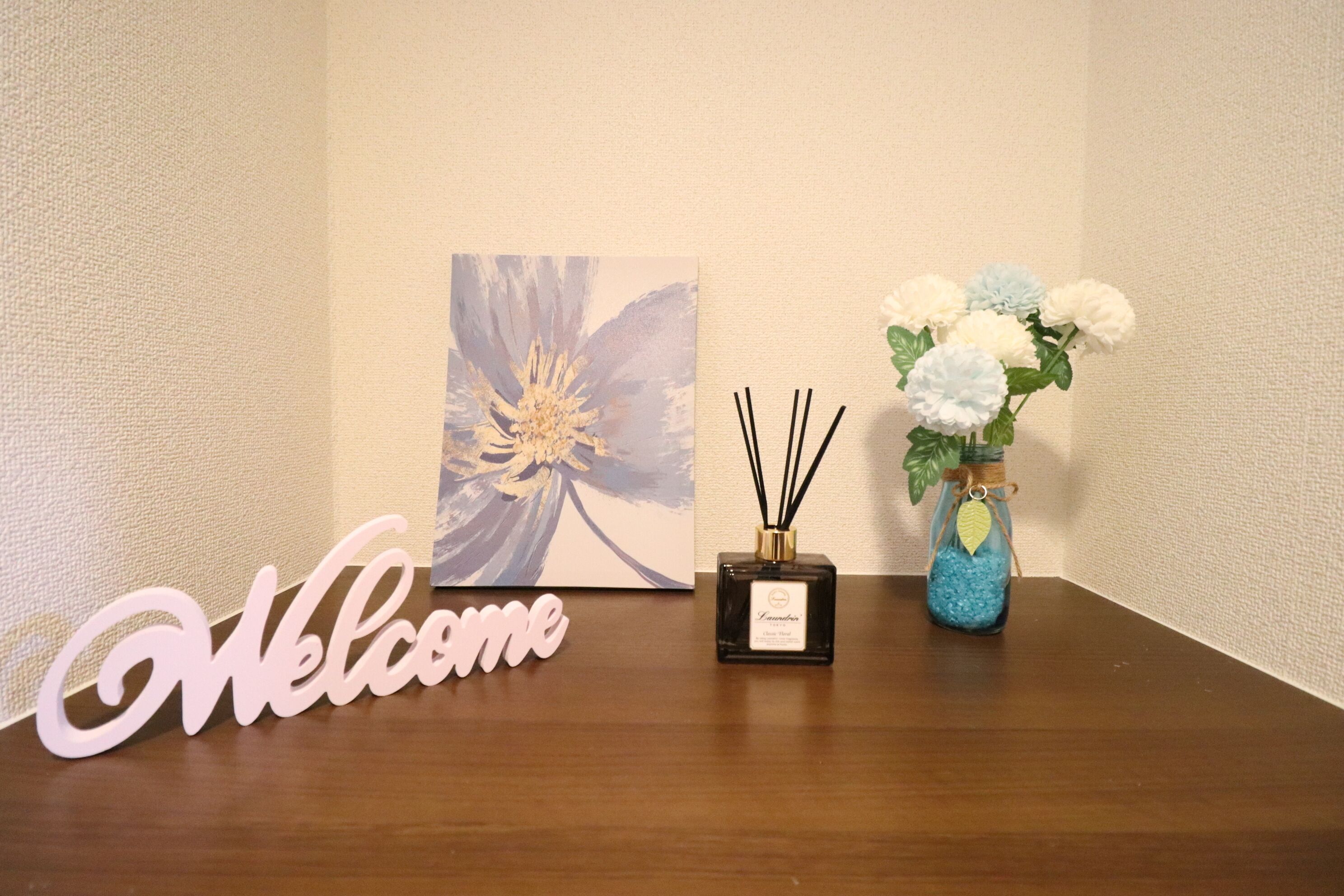 A wooden table with a 'Welcome' sign, a framed artwork, a small black bottle, and a blue vase with flowers.