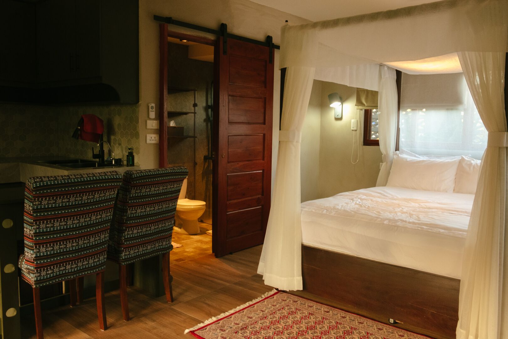 A bedroom with a bed under a canopy, a wooden door, a chair, and a small table with a lamp.