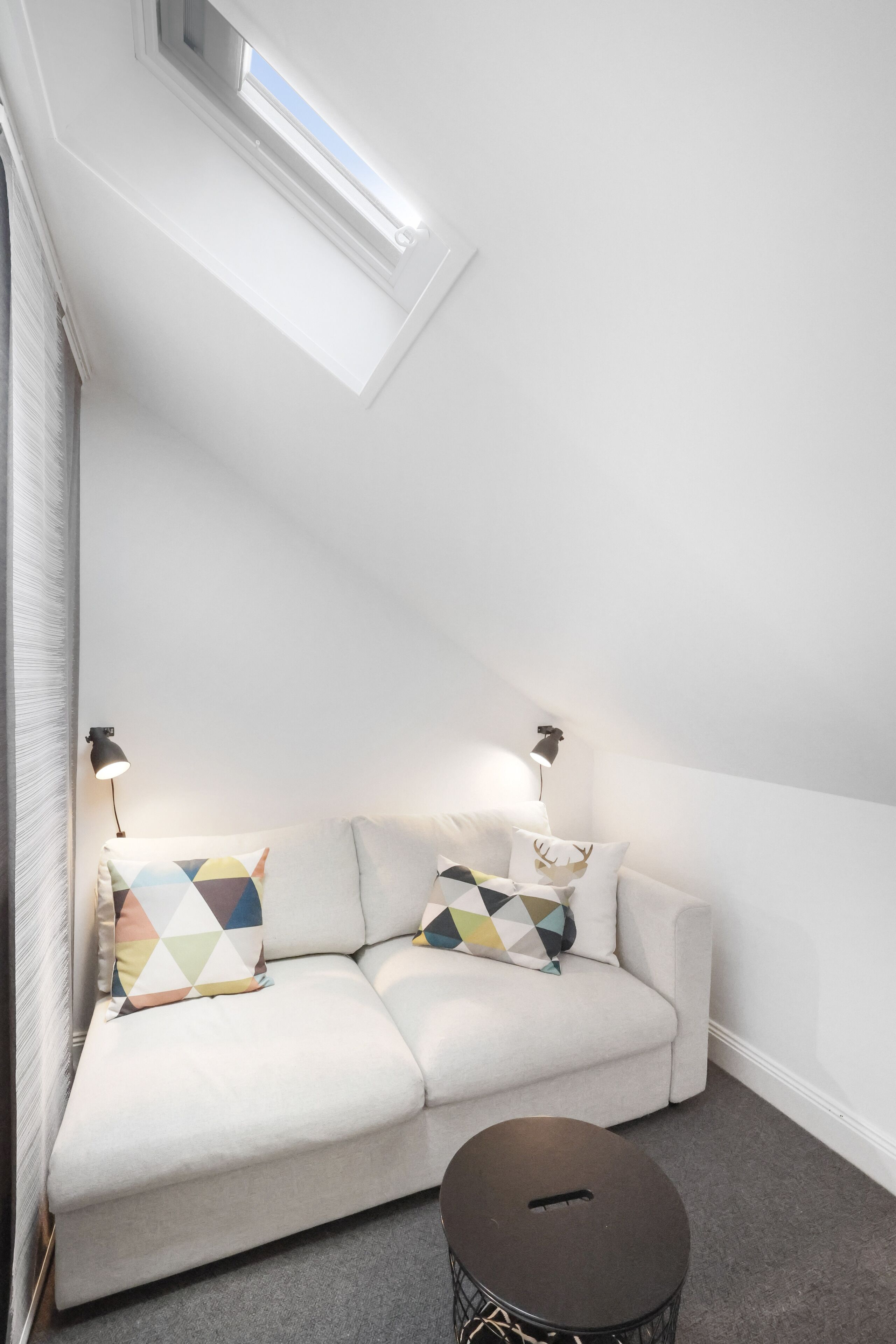 Ett modernt sovrum i loftstil med en vit soffa, mönstrade kuddar, ett runt soffbord och ett takfönster.