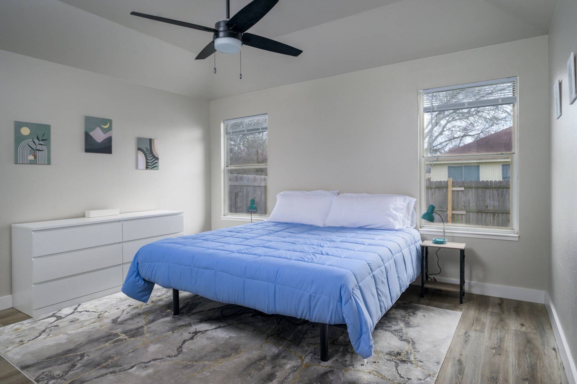 A bedroom with a bed, a ceiling fan, a nightstand, a desk, and two windows.