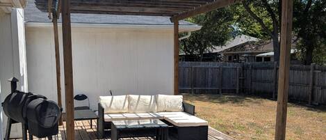 A covered outdoor patio with a grill, a cushioned seating area, and a wooden deck.