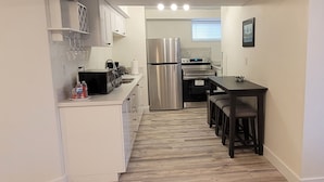 A compact kitchen with white cabinets, a stainless steel refrigerator, and a dining area with a dark table and chairs. - Cozy Home Away from Home (Kingston)