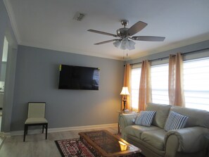 Uma sala de estar com ventilador de teto, televisão de tela plana na parede, sofá com almofadas listradas, uma poltrona e uma mesa de centro.