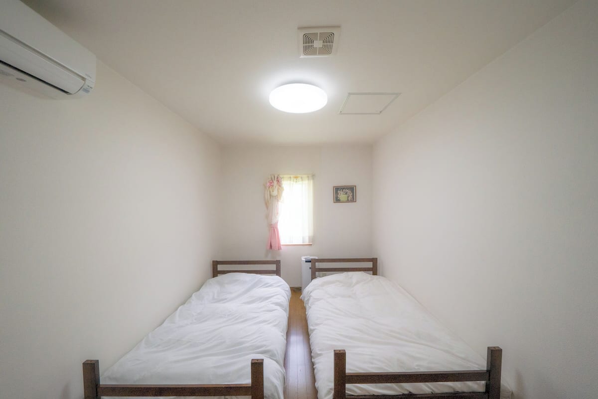 A narrow room with two single beds, a ceiling light, a window with a curtain, and a wall-mounted air conditioner.