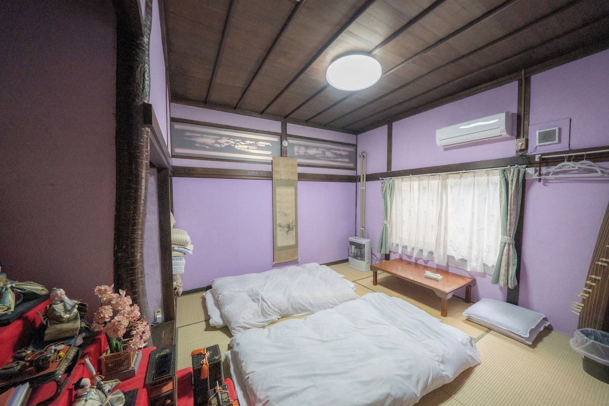 A room with two beds, a small table, an air conditioning unit, and a window with curtains.