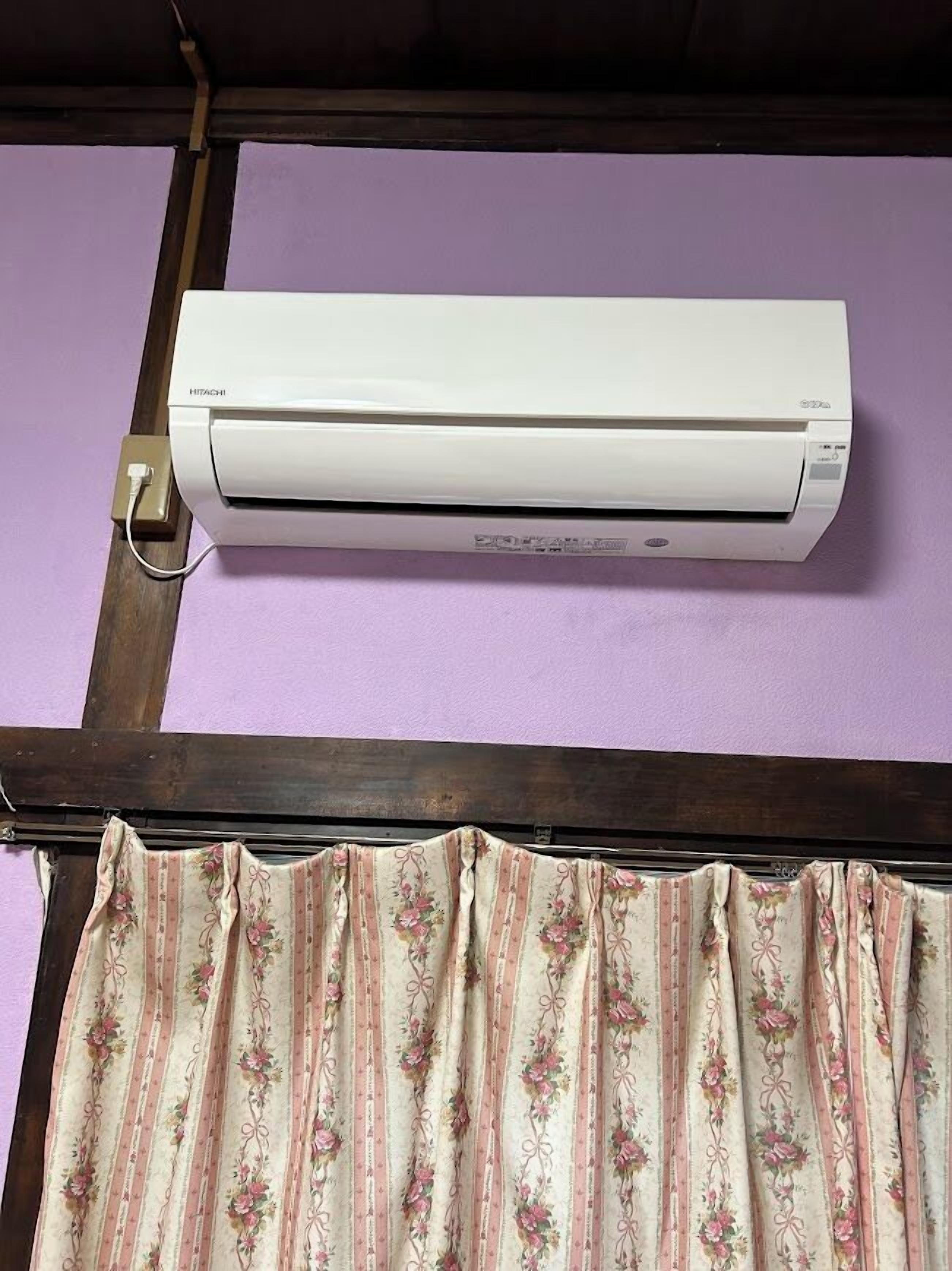 A white air conditioner mounted on a purple wall, with a wooden beam above and a floral curtain below.