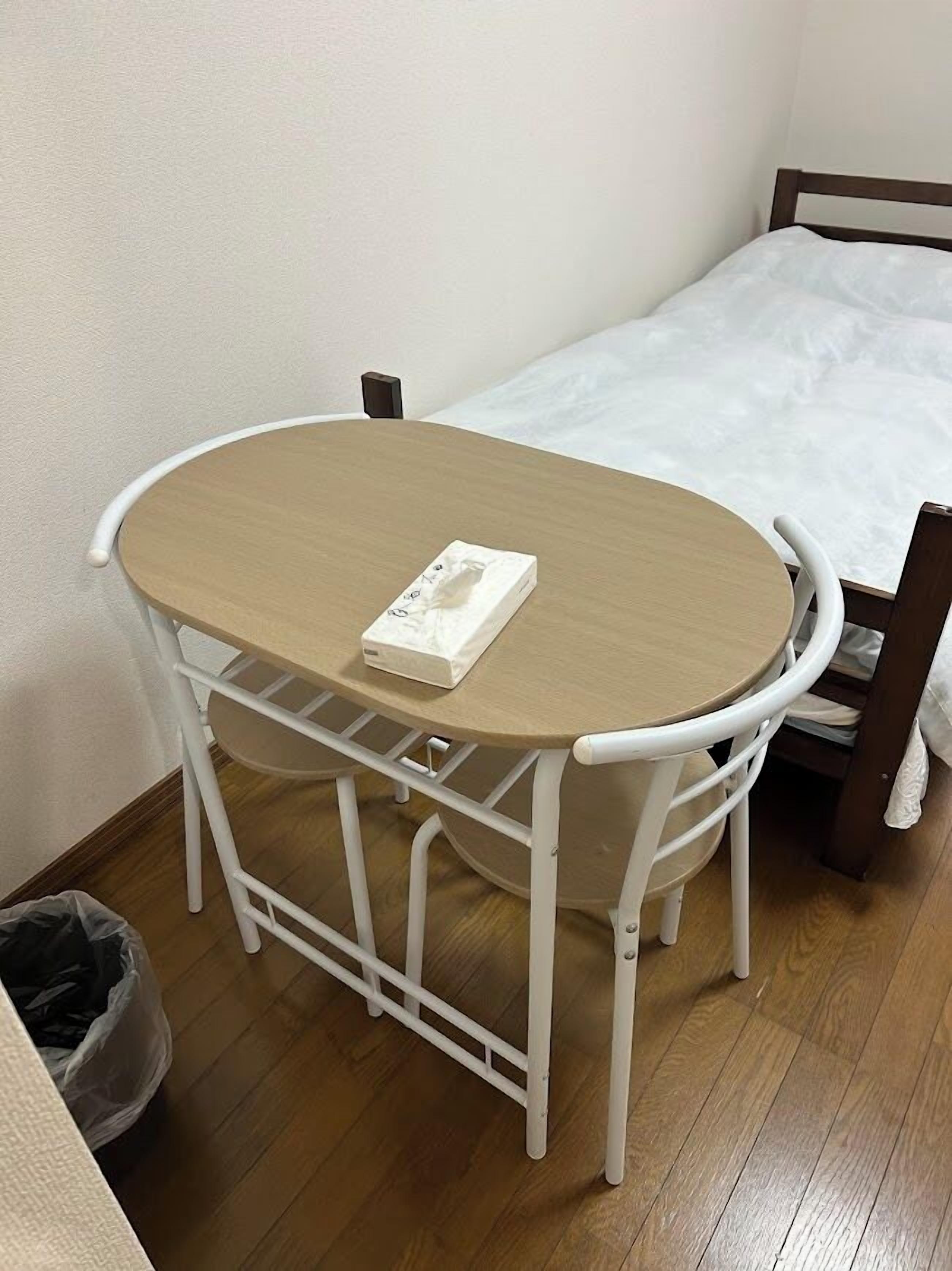 A small, single-room accommodation with a wooden floor, a bed with white bedding, a white metal table with a single chair, and a small trash bin.