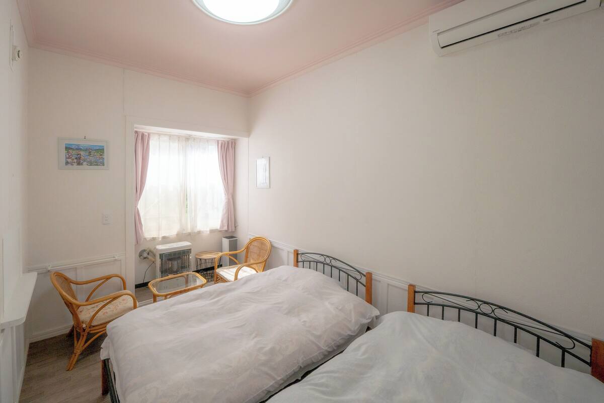 A bedroom with two beds, a window with curtains, a small table, and a chair.