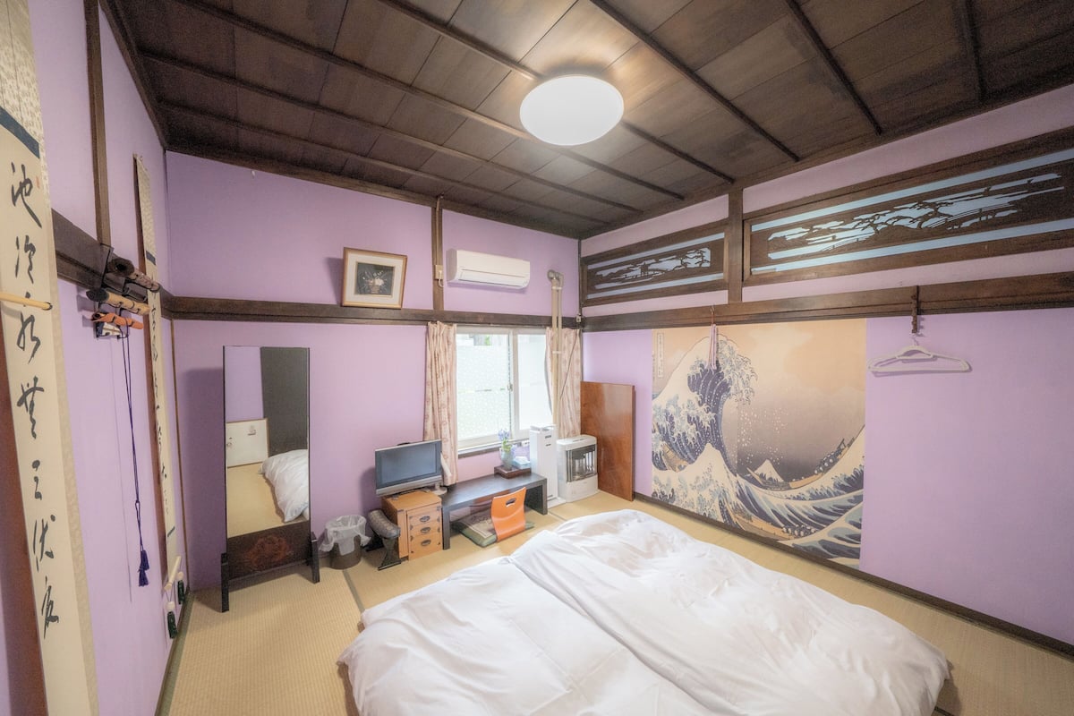 A room with a bed, a desk with a computer, a mirror, and a wall with a Japanese artwork.