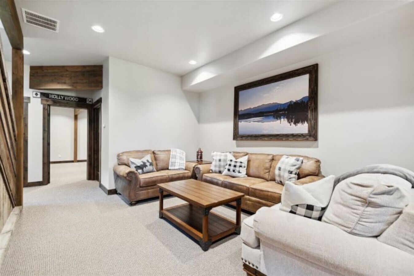 A living room with a brown leather sofa, a wooden coffee table, and a white sofa. There is a painting of a lake on the wall.