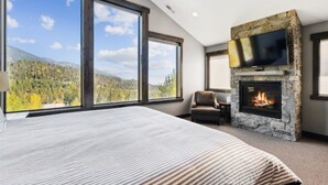 Une chambre avec un grand lit, une cheminée en pierre, un téléviseur fixé au-dessus, et une vue sur une forêt à travers de grandes fenêtres.