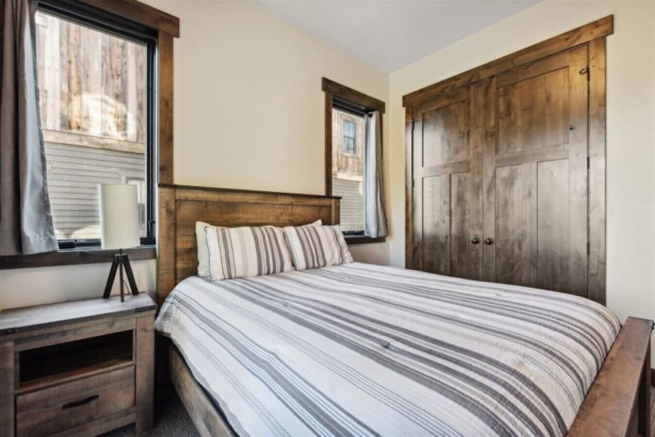 A bedroom with a large bed, wooden headboard, two windows with blinds, a wooden wardrobe, and a bedside table with a lamp.