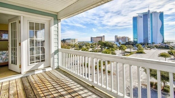 A wooden deck with a white railing, a glass door, and a view of a cityscape including a tall building. - Ocean Views New 4BR Steps from Beach (Myrtle Beach)