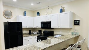 A kitchen with white cabinets, a black refrigerator, a microwave, and a granite countertop. There are two white bar stools with wooden seats. - Ocean Views New 4BR Steps from Beach (Myrtle Beach)
