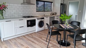 A modern kitchen with white cabinetry, a built-in oven, a washing machine, and a dining area with a round table and black chairs. - Roberts Beach Escape in Tairua is 3-5 minute walk from the beach (Tairua)